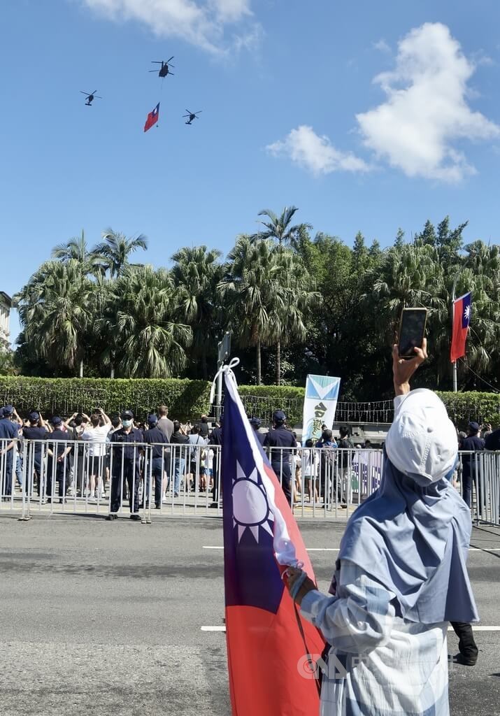 Attendees of the National Day celebrations raise their phones to the sky while a CH-47SD Chinook flies the nation's flag across the sky, chaperoned by two UH-60M Black Hawk helicopters. CNA photo Oct. 10, 2025
