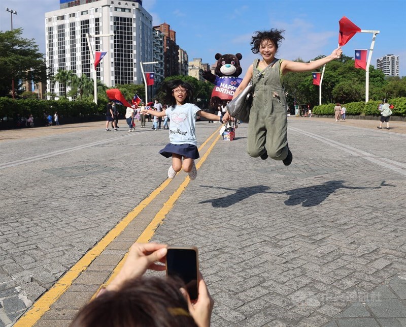 Pemerintah Kota Taipei mengadakan upacara pengibaran bendera dan perayaan di Plaza Gedung Kantor Kepresidenan pada tanggal 10 Oktober untuk memperingati ulang tahun ke-115 Republik Tiongkok. Orang tua dan anak-anak berpose untuk foto bersama maskot pemerintah kota, "Beruang Zan." (Sumber foto: CNA, 10 Oktober 2026)