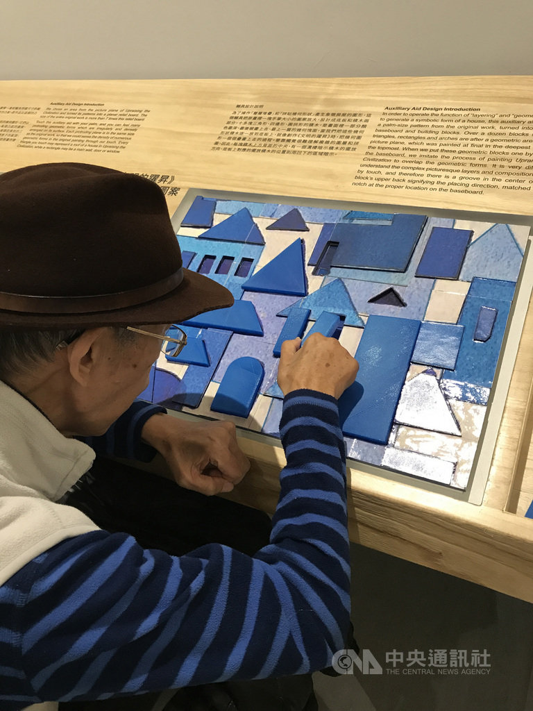 國美館為畫家張永村的水彩畫作「文明的躍昇」打造輔具,有10多塊三角形、四邊型、圓拱型積木,是原畫中一部分顏色最深、最後被畫上去、最上一層的幾何塊面。已2度中風的張永村(背對者)親身體驗,藝術輔具也是復健小幫手。(國美館提供)中央社記者陳偉婷傳真 109年1月29日