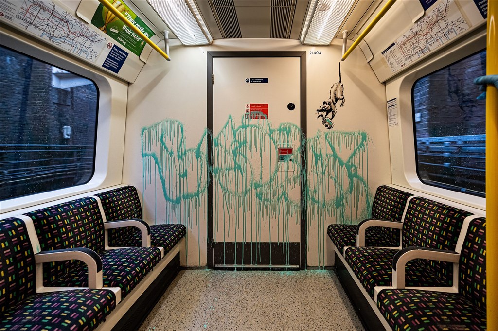英國知名塗鴉藝術家班克西受疫情啟發,在倫敦地鐵環線車廂內作畫,並留下他的大名,旁邊是隻老鼠用尾巴吊掛在握桿上,手裡拿著消毒液。(圖取自班克西官方網頁banksy.co.uk)