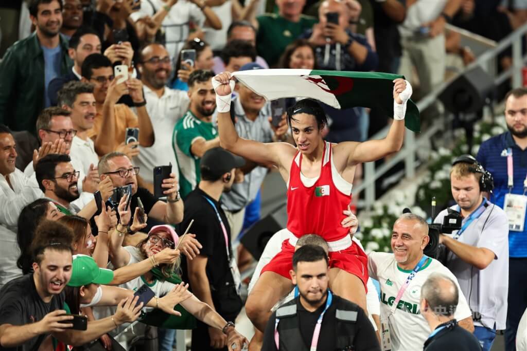 在2024巴黎奧運捲入性別爭議的阿爾及利亞女拳手克莉芙（Imane Khelif）（紅衣）法國時間9日晚間在女子66公斤級金牌戰以5比0完勝奪金， ​​​​​​​實踐賽前盼讓阿爾及利亞與整個阿拉伯世界驕傲的心願，賽後她開心坐在團隊成員肩頭、手持國旗開心繞場。（圖／中央社）