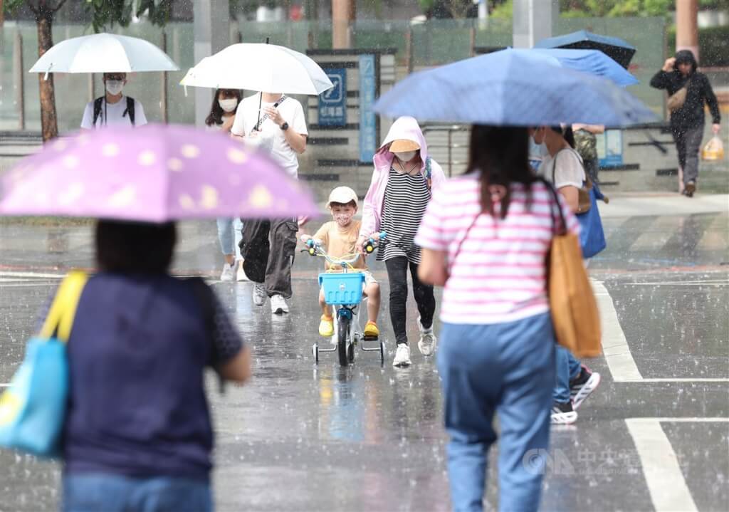 台北市信義區午後下起雷陣雨，民眾撐起雨傘遮擋雨勢，有家長趁雨勢漸歇，牽著騎腳踏車的小孩過馬路。（圖／中央社）