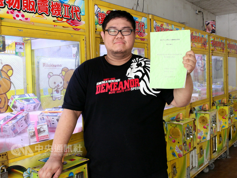 中正大學成人及繼續教育系碩士研究生趙亮鈞以「夾娃娃」當作論文主題,展開研究訪談,更在寫作期間陸續開設3家夾娃娃機店面;論文完成後,趙亮鈞開心拿著論文在機台前合影。(中正大學提供)中央社記者江俊亮傳真 107年8月8日