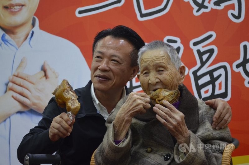 嘉義縣補助長者做活動假牙，縣府14日請來百歲人瑞陳周玉緞（右）啃雞腿見證成效，未裝假牙前她無法正常進食，身體虛弱無法行走，目前恢復健康，能用助行器走動。左為嘉義縣長翁章梁。中央社記者蔡智明攝　109年12月14日