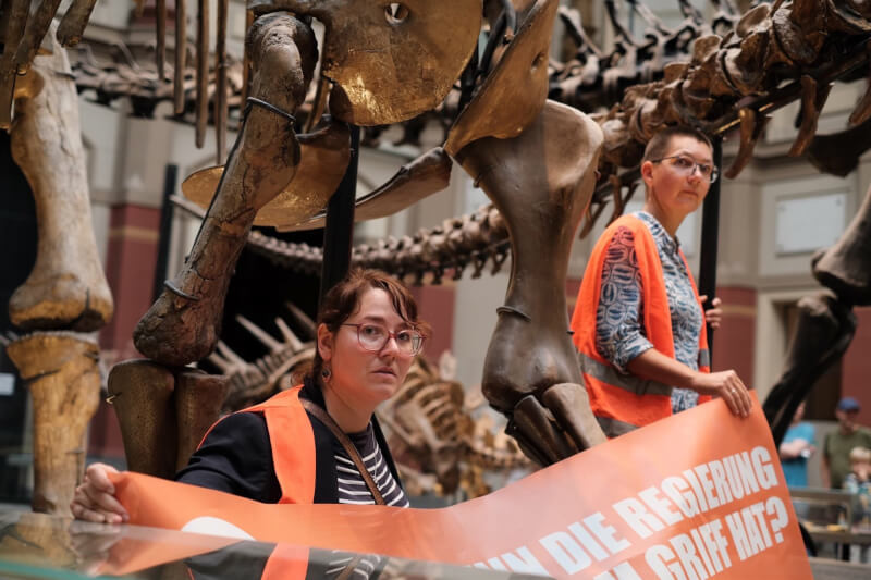 德國兩名環保人士30日在柏林自然博物館將自己黏在恐龍骨架下方的支柱，抗議政府氣候政策。（圖取自twitter.com/AufstandLastGen）