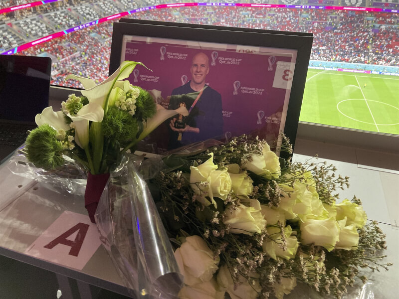 美國體育記者華爾在卡達報導世界盃足球賽過世後，國際足球總會（FIFA）在原本預留給他的記者座位擺放照片鮮花悼念。（美聯社）