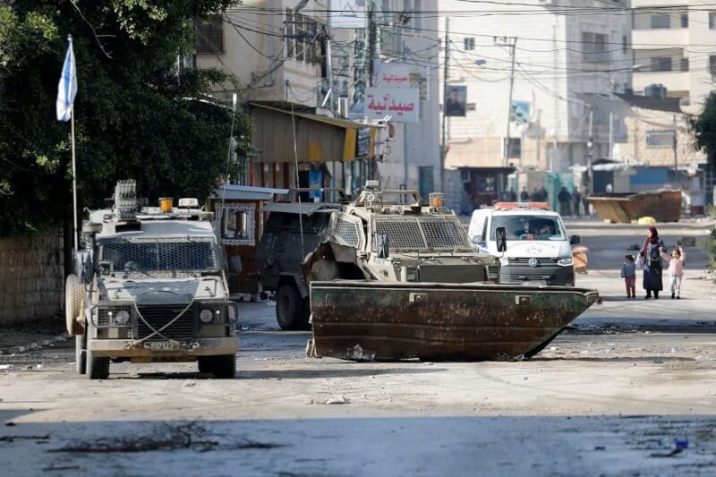 以色列突襲約旦河西岸城市哲寧，一名婦女14日帶著孩子行經以軍戰車旁。（路透社）