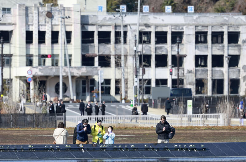 日本民眾11日雙手合十悼念311大地震，後方是地震遺址石卷市門脅小學。（共同社）