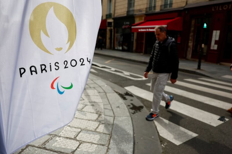 圖為一名男子3月25日在法國街頭經過巴黎奧運旗幟旁。（路透社）