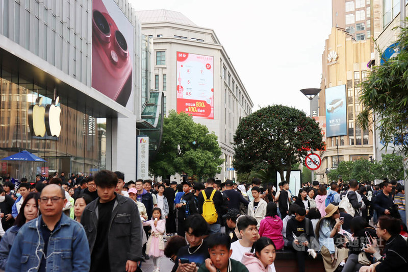 上海南京路步行街在「五一」假期首日出现人潮，这条路两旁有各种吃喝玩乐等消费服务，知名电子消费品牌苹果和华为的大型店也在此。图中，这两家巨头隔街相望，中间出现的看板广告，则是上海市政府在5、6月力推的购物节活动。中央社记者张淑伶上海摄  113年5月1日