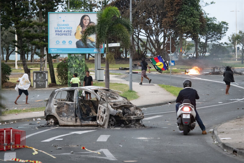 新喀里多尼亞發生暴動，14日街頭可見燒毀的車輛，一名男子舉著卡納克社會主義民族解放陣線的旗幟。（法新社）