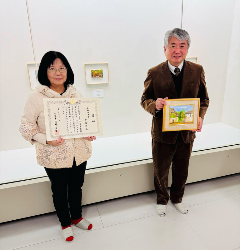 來自台灣的創作者李麗琴(左)以油畫加上刺繡完成明信片,獲得日本車屋美術館主辦「10x15世界大賽」最高榮譽市長獎,她11日到日本栃木縣小山市領獎,並以車屋美術館外觀「小川家住宅」為主題創作畫作致贈館方,由小山市長淺野正富(右)代表接受。(李麗琴提供)中央社記者邱祖胤傳真 114年1月11日