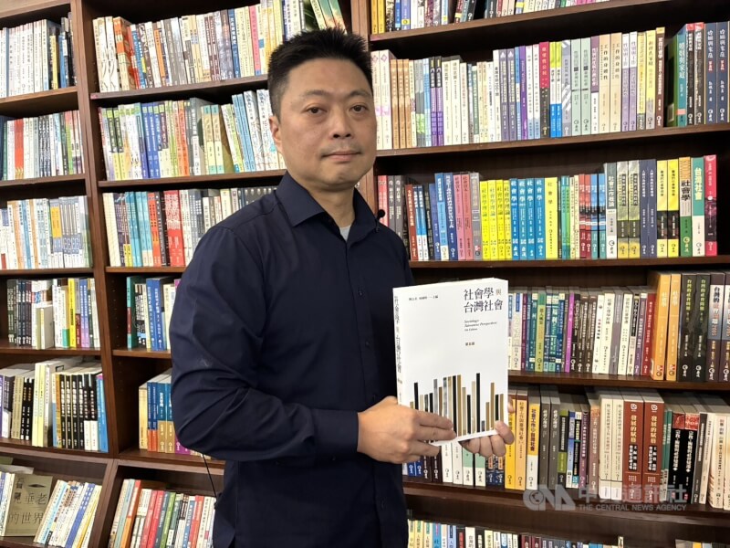 麗文總經理楊宏文拿著旗下品牌巨流出版的「社會學與台灣社會」,為台灣社會學系學生必讀書目,至今已出到第5版,他身後則是擺滿麗文出版品的書牆。中央社記者陳政偉攝 114年10月9日