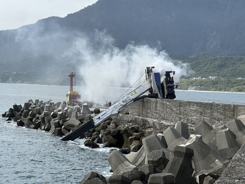 台東成功漁港30日中午一輛吊車在碼頭吊掛消波塊時翻覆，車上其中1人在駕駛座已明顯死亡，另1人腿部遭車體壓住受困，消防人員救出後送醫救治，吊車還冒煙起火，隨即被警消撲滅。（民眾提供）中央社記者李先鳳傳真　114年11月30日