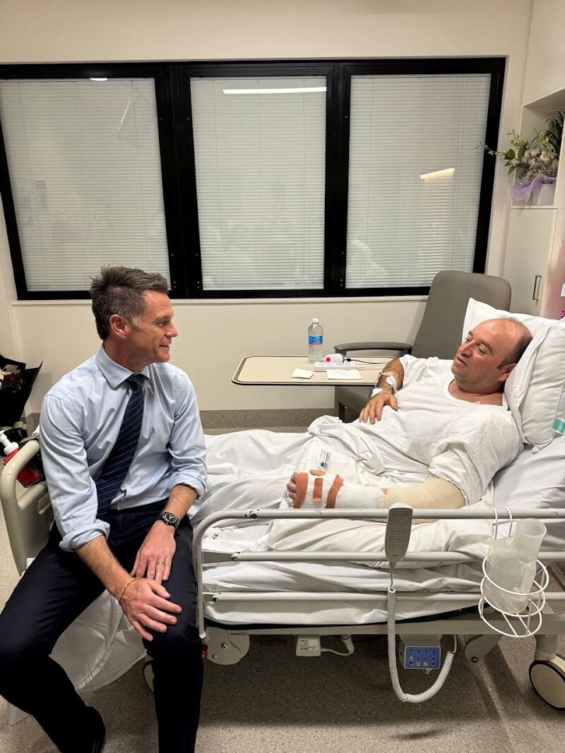 澳洲新南威爾斯州長柯民思（左）15日前往醫院探視徒手奪槍制伏歹徒的菸草專賣店老闆阿邁德（右）。（圖取自x.com/ChrisMinnsMP）