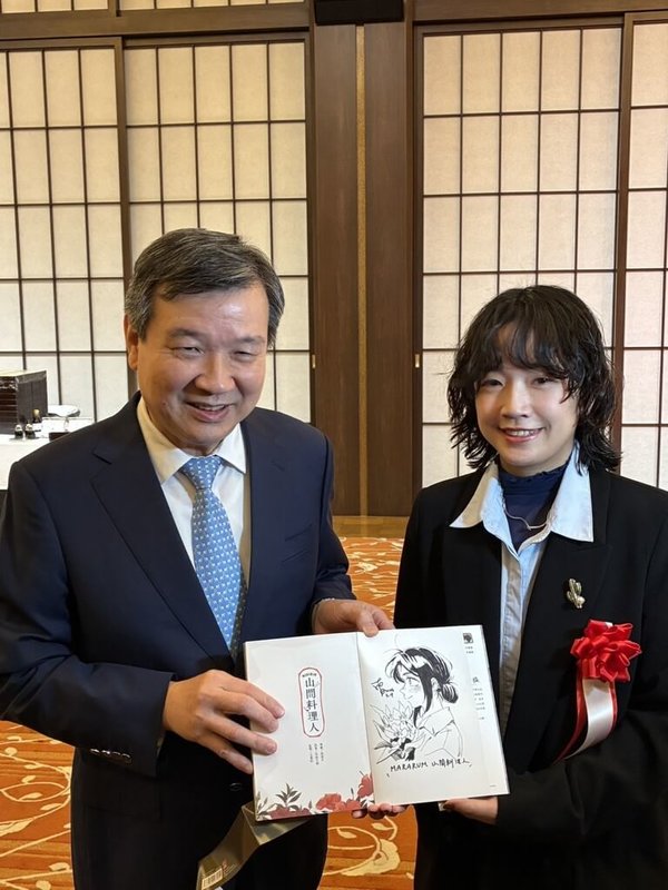 日本外務省主辦的第19屆日本國際漫畫獎頒獎典禮4日在東京飯倉公館舉行。台灣漫畫家南南日（右）以作品「Mararum：山間料理人」榮獲優秀獎（銀獎），致贈得獎作品予駐日代表李逸洋，並於扉頁親筆繪上主角「巴奈」畫像留念。（台灣文化中心提供）中央社記者戴雅真東京傳真 115年3月4日