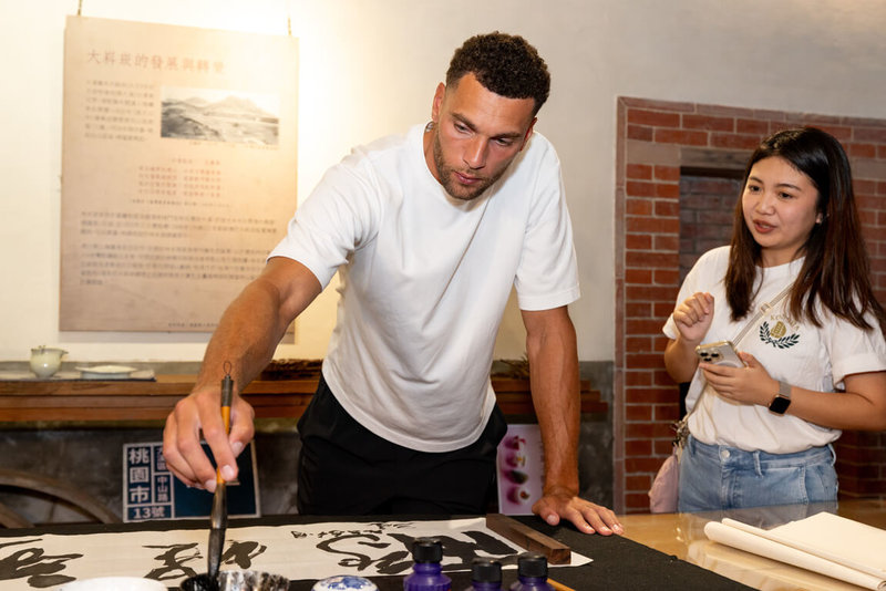 美國職籃NBA球星拉文（Zach LaVine）（左）2日走訪大溪老街，觀賞書法老師揮毫「鵬程萬里」，呼應他2度奪得NBA灌籃大賽冠軍的飛人形象，拉文親自在作品落款，也提筆寫下草書「飛」字留念。（桃園市政府觀光旅遊局提供）中央社記者吳睿騏桃園傳真　115年8月2日