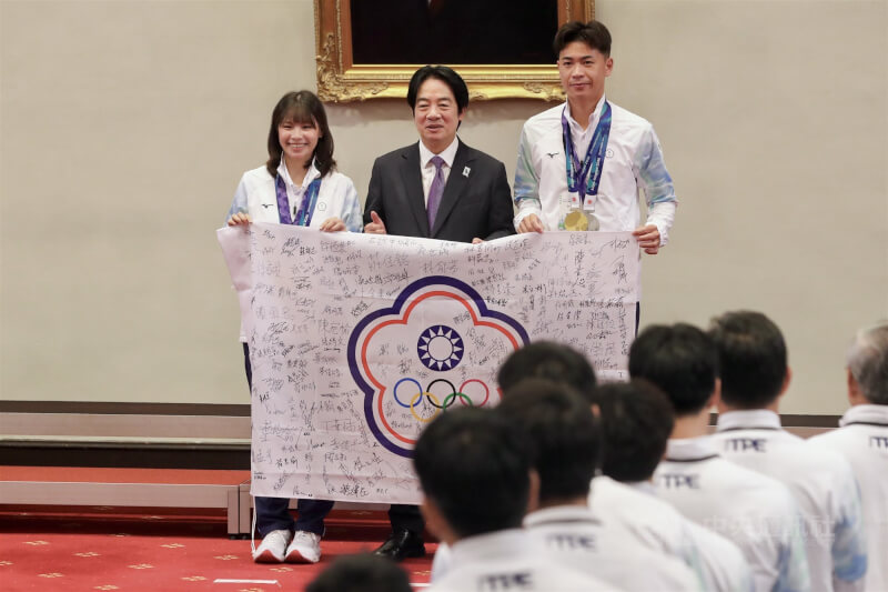 總統賴清德（中）6日在總統府接見我國參加「2026年第20屆愛知名古屋亞洲運動會」代表團，亞運金牌軟式網球混雙選手余凱文（右）、黃詩媛（左）代表致贈賴總統參賽選手簽名會旗。中央社記者裴禛攝 115年10月6日