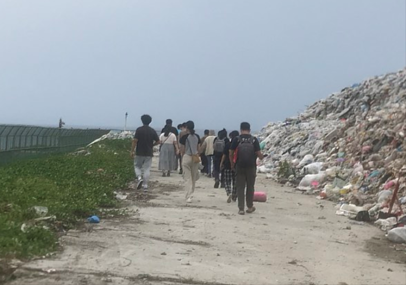 師生走在垃圾運送道上，右邊是高高的垃圾山，左邊圍籬後面就是海。