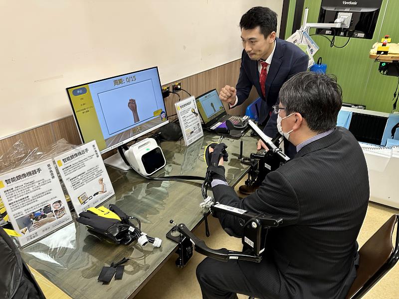 日本帝京平城大學鎗田英樹教授(右),在亞大附醫智能復健機器人中心,體驗訓練手功能機器。