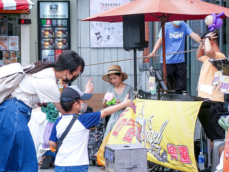 老街上，一位媽媽牽著孩子停下腳步，靜靜聆聽著樂團的演奏。儘管下著雨，仍用行動傳遞鼓勵與肯定，那一份溫柔與真心，就像一把傘，撐住了孩子們努力發光的夢。