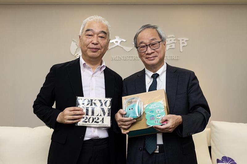 文化部長李遠(右)致贈「臺灣文化in大阪.關西世博」吉祥物「a-We」痛包及徽章等文宣品予東京影展資深策展人石坂健治(左),並說明「a-We」多元、包容、韌性的形象。