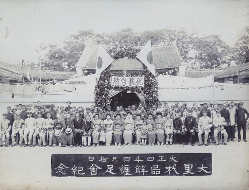 臺中州廳長官私人寫真帖（二）55 大里杙街在今臺中市東南部，1915年4月該街放足者已達1,200人，未放足者僅餘152人。大里杙解纏足會乃於4月10日上午11時集合已放足會員300人，在大里杙區役場前舉行解纏足祝賀會，會後舉行餐會，並贈送出席會員木屐一雙。本照片係祝賀會結束後所拍攝紀念照，照片中臨時搭建牌坊上方寫著「祝我自然」4字。