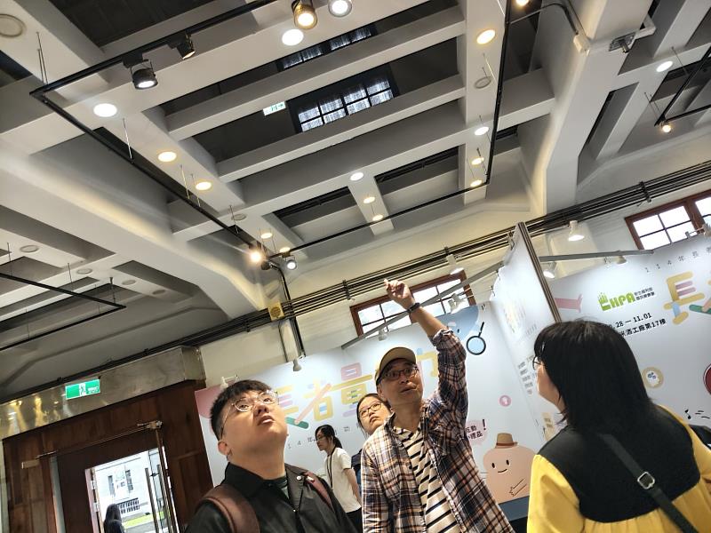 老師帶領學生觀察舊酒廠修復木造房時增添的鋼骨結構，背景為廠商舉辦展覽。