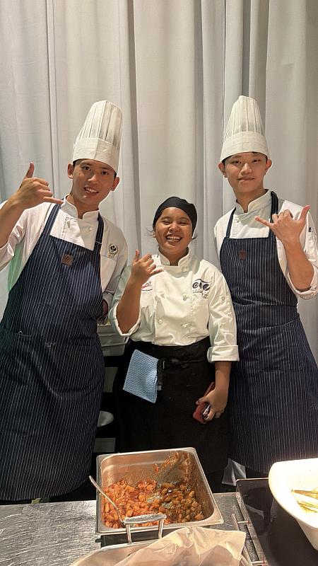 高餐大西廚系學生與關島社區學院學生開心分享廚藝製作經驗。依序為西餐廚藝系劉宸睿（左1）、陳韋誠（右1）、關島社區學院學生（中）。