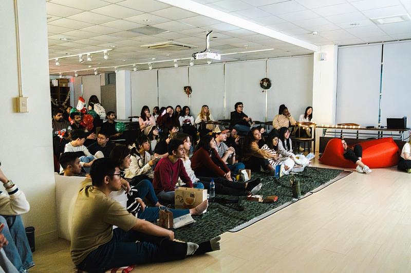 學生們圍坐在鋪滿地毯、抱枕與坐墊的舒適空間，一邊觀賞聖誕電影，一邊享受療癒而愉悅的節日氛圍。