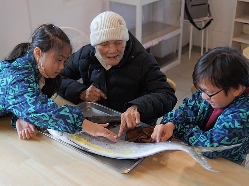 一年級周同學鼓勵林阿嬤觸摸鬼頭刀，六年級周王同學解說魚的特徵。