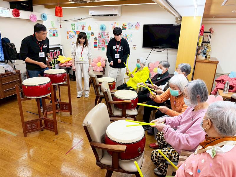 音樂系盧煥韋教授帶領師生,手持響鼓、手鑼等樂器走入日照中心。在《望春風》等經典旋律帶動下,長者重拾笑容並隨之敲擊律動,展現藝術療癒在維持認知功能與改善情緒上的輔助價值。