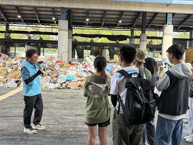 吳隊長說明垃圾回收作業，遠處有清潔隊員站在輸送帶後方，做垃圾分類。