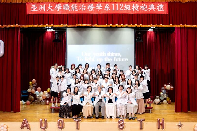 亞大職治系授袍典禮舞台大合照，師生身穿白袍溫馨比心，共慶專業傳承。