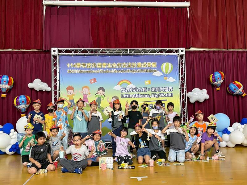 參與「114學年度外國學生合作交流計畫成果展」的學童於活動舞台前開心合影，手持各國文化體驗活動完成後獲得的小禮物與作品，展現豐碩學習成果與跨文化探索的喜悅。