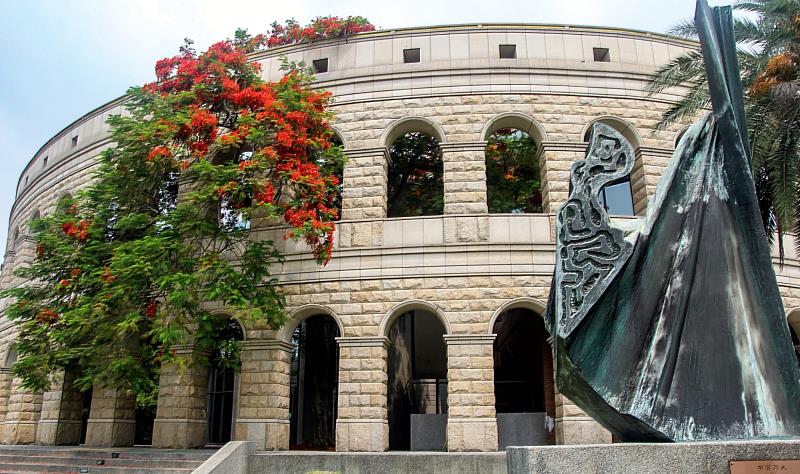 亞大體育館羅馬式建築旁,火紅鳳凰花熱情盛開,與青銅雕塑交織出初夏迷人的校園風光。