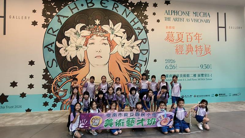 臺南市麻豆國民小學美術藝才班師生於「慕夏百年經典特展」主視覺牆前留下大合照。/麻豆國小提供