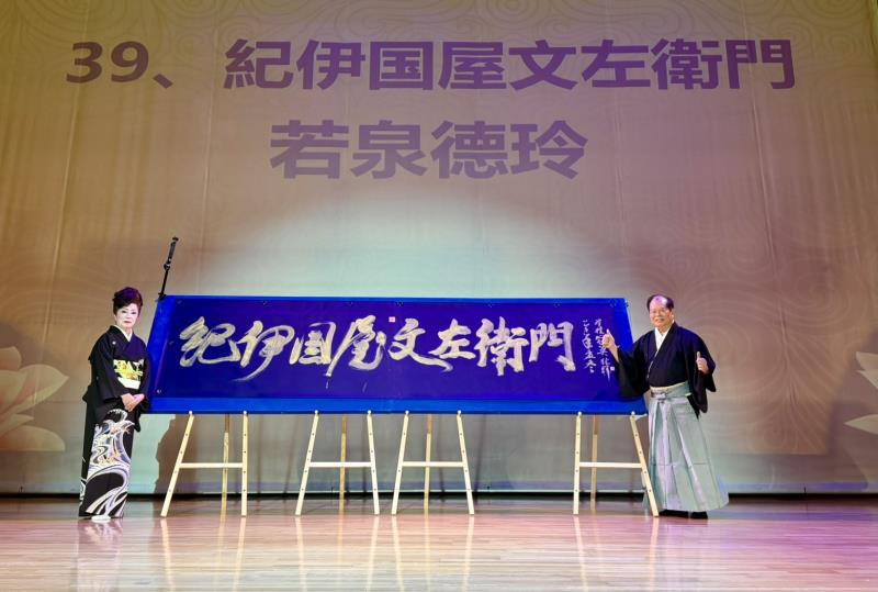新邦樂舞踊若泉流台灣分家家元若泉德玲（林汶玲老師，左）與林暉博士（右）於舞台上共同展示林博士現場揮毫書法巨作〈紀伊國屋文左衛門〉。作品氣韻生動、筆勢雄健，象徵日台藝術文化的交融與友誼長存。圖片提供 必紅演藝